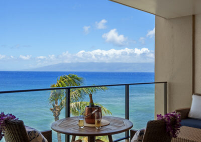 Bright blue sweeping ocean view from patio with outdoor chairs and champagne in ice bucket with palm tree blowing in breeze