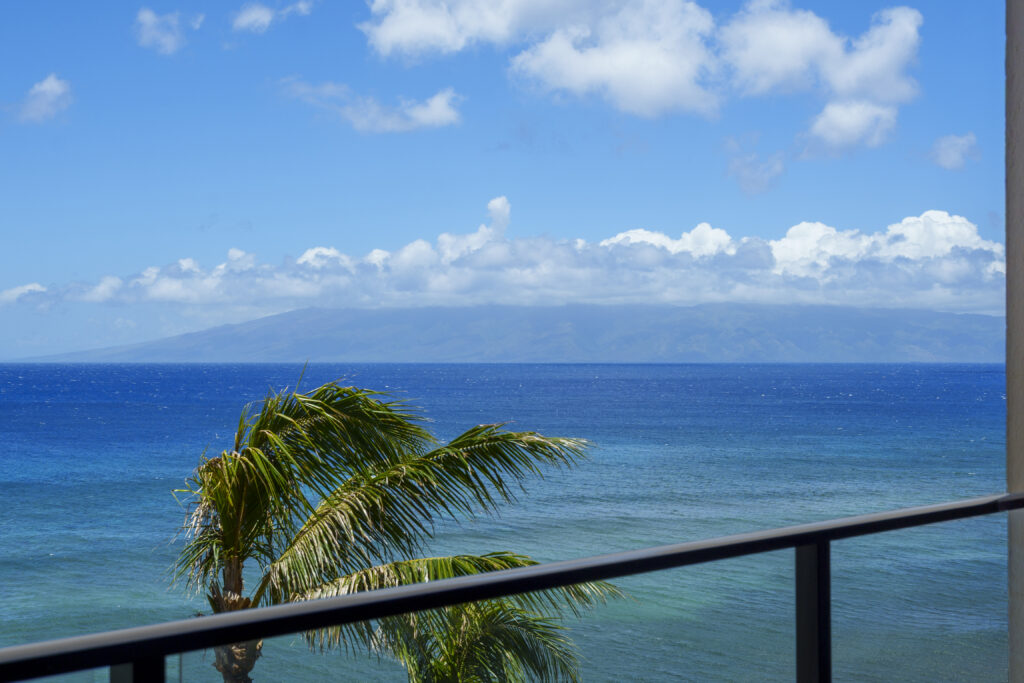 Bright blue sweeping ocean view with outer island of Molokaiin distance and palm tree leaves in waving with breeze.