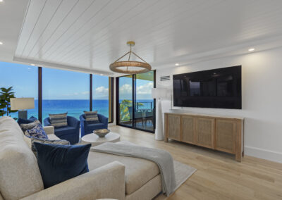Bright modern living room with off-white sectional and blue chairs - large wall-mounted TV and floor to ceiling windows overlooking ocean