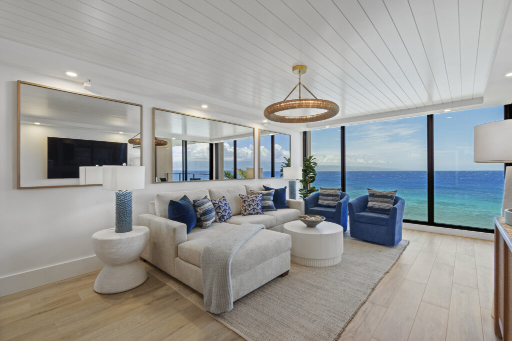 Bright modern living room with off-white sectional and blue chairs - mirrored back wall and floor to ceiling windows overlooking ocean