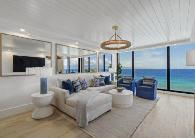 Bright modern living room with off-white sectional and blue chairs - mirrored back wall and floor to ceiling windows overlooking ocean